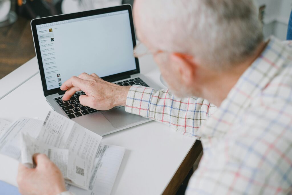 Elderly man at desk handling finances online with documents and laptop.